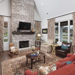 A cozy living room with a brick fireplace, mounted TV, and patterned curtains. There's a red sofa, armchairs, and a wooden rocking chair. A rug and coffee table complete the space.