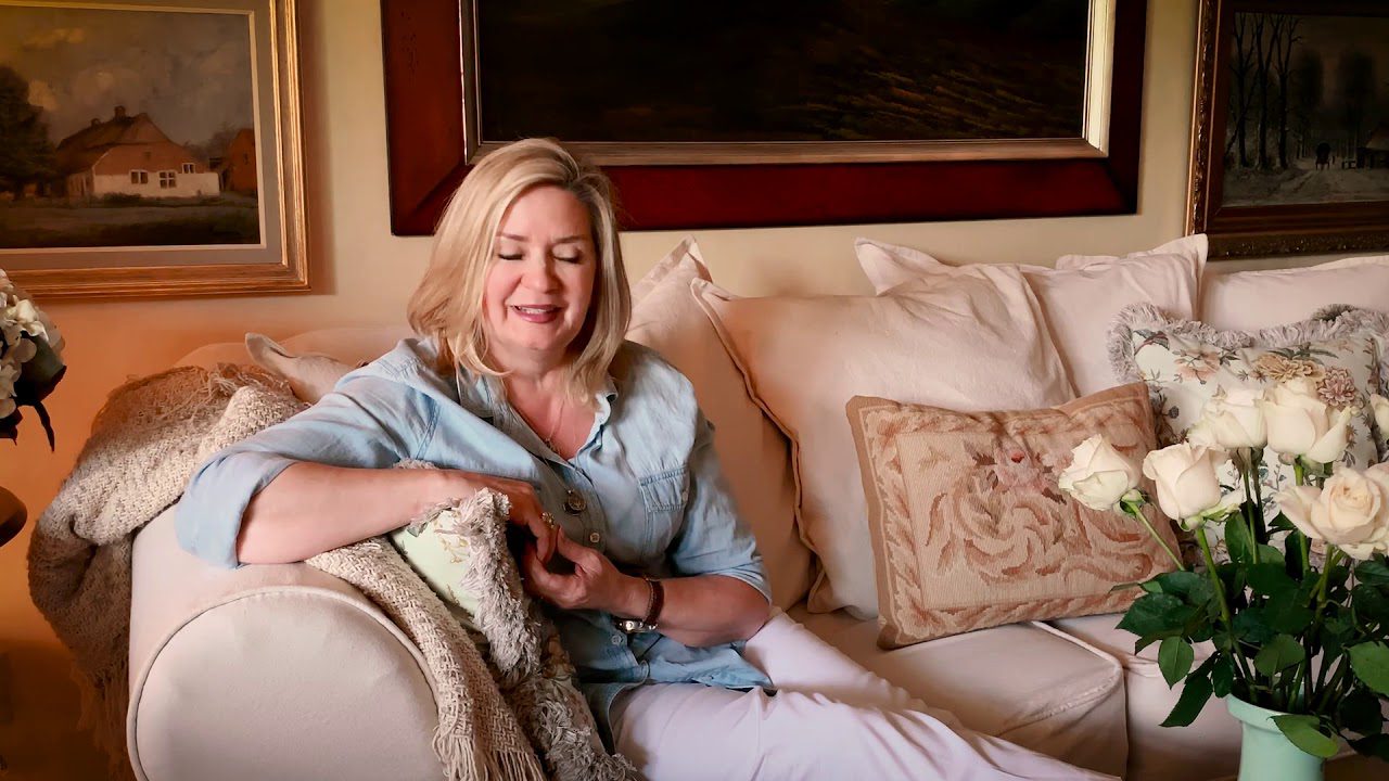 A woman sitting on a beige sofa in a living room, surrounded by cushions and a bouquet of white roses on a coffee table. Framed artwork is visible on the walls.