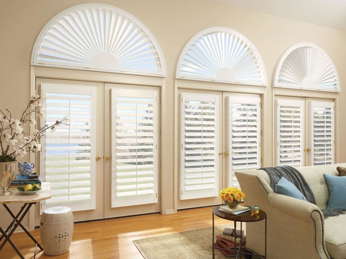 A living room with three French doors, arched transom windows, and white shutters is adorned with elegant shades. The cozy space features a sofa next to a side table with a vase and a ceramic stool on light wood flooring.