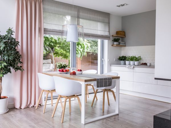 Modern dining area with a wooden table, white chairs, and a hanging light. Large windows feature Roman shades with pink accents, offering views of greenery outside. White cabinets and plants tastefully decorate the space.