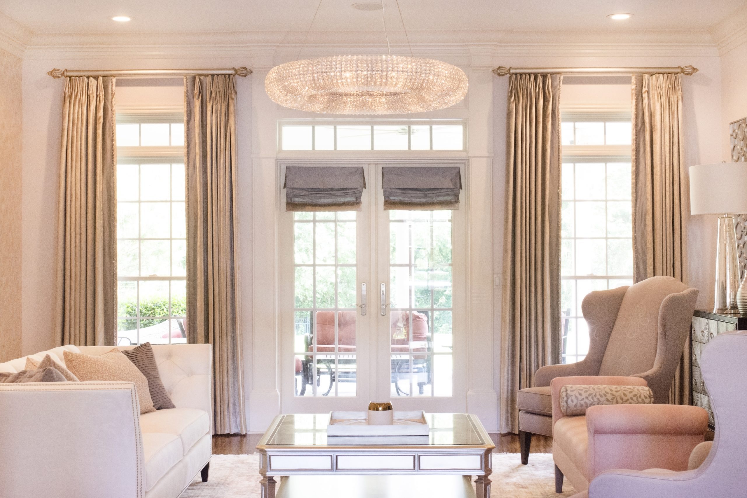 Elegant living room with a chandelier, large windows adorned with custom window treatments, beige curtains, a white couch, armchairs, and a mirrored coffee table.