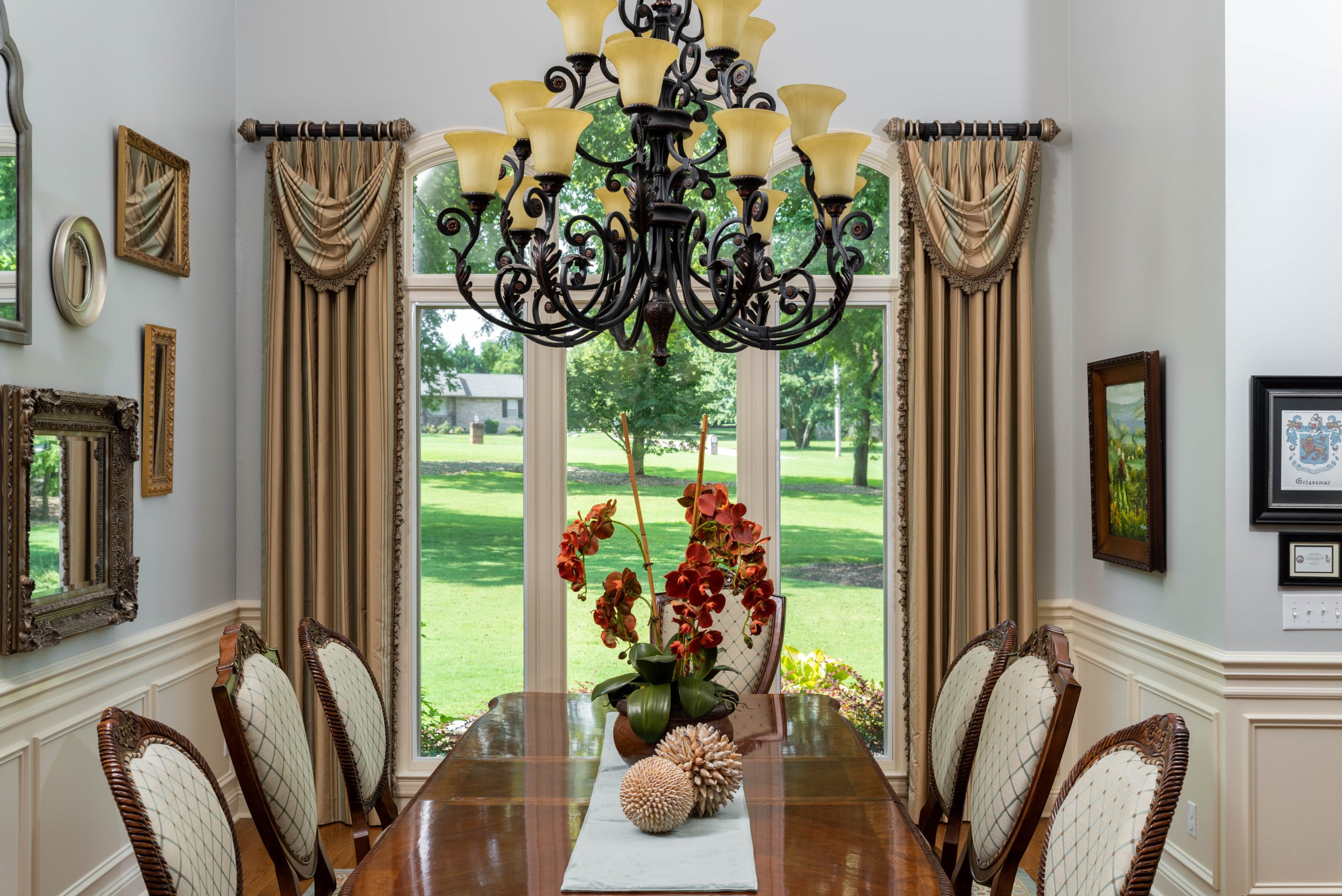 Dining room with a long wooden table, chairs, a central floral arrangement, ornate chandelier, large windows adorned with elegant draperies, and framed wall art, overlooking a green outdoor area.