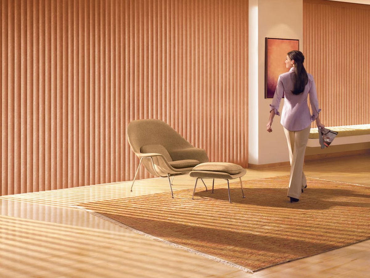 A woman strolls through a room with ribbed walls and window blinds filtering soft light onto a patterned rug. She is wearing a light purple blouse and beige pants, accompanied by a modern chair and ottoman.