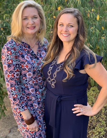 Two women are smiling outdoors, standing side by side. One wears a patterned dress, the other a navy dress with a necklace. Greenery frames them like lush Chattanooga window treatments gently swaying in the background.