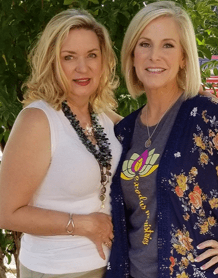 Two women stand together outdoors, their smiles as vibrant as the floral prints they wear. One wears a white top and green necklace; the other sports a floral cardigan over a lotus-design tee, with trees gently framing their moment like delicate Chattanooga window treatments.