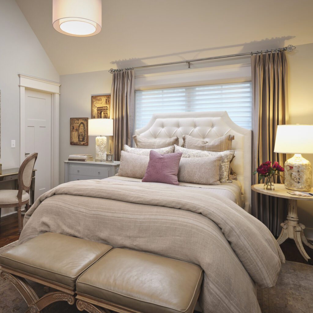 Elegant bedroom with a large bed, tufted headboard, and layered bedding. Two lamps on side tables, a bench at the foot of the bed, a desk to the left, and decorative art on the walls.