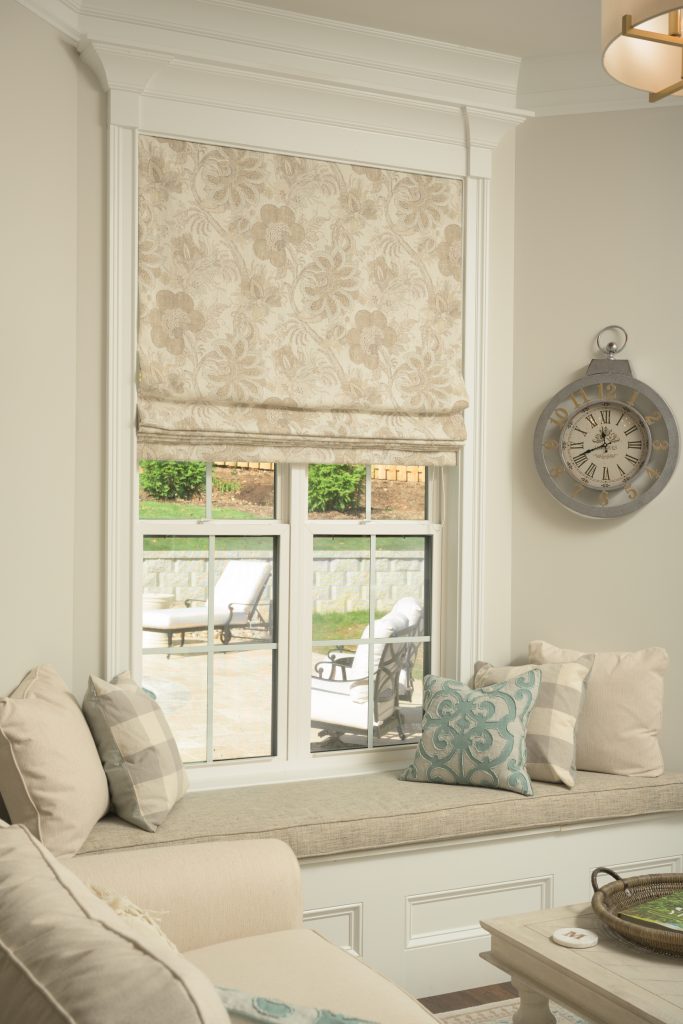 A cozy nook features a window with patterned Roman shades, a built-in bench seat with cushions, beige walls, and a decorative wall clock. Outdoor patio furniture is visible through the window.