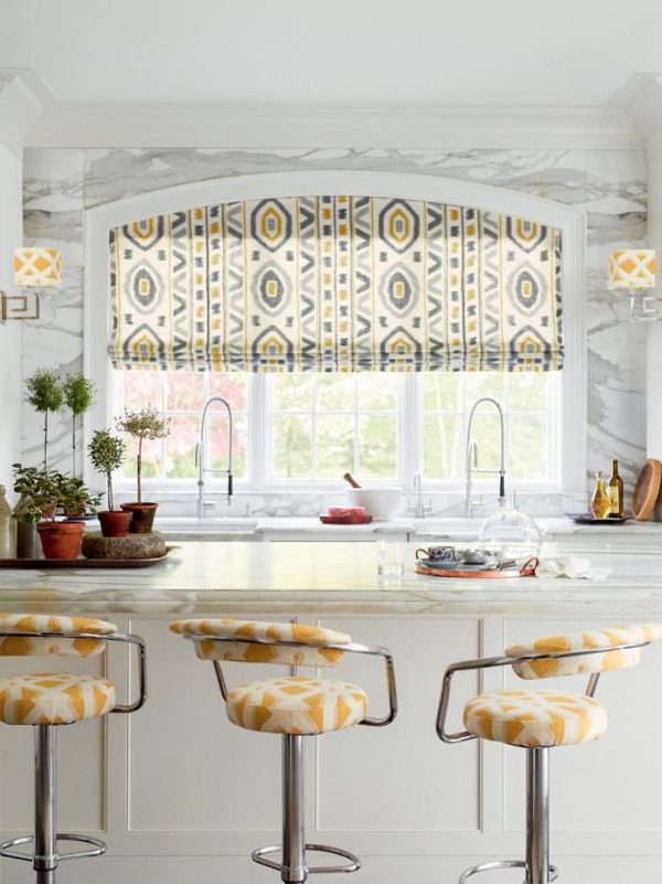A kitchen with stylish Roman shades as window treatments above the sink, decorative plants on the counter, and three stools with matching patterned cushions.