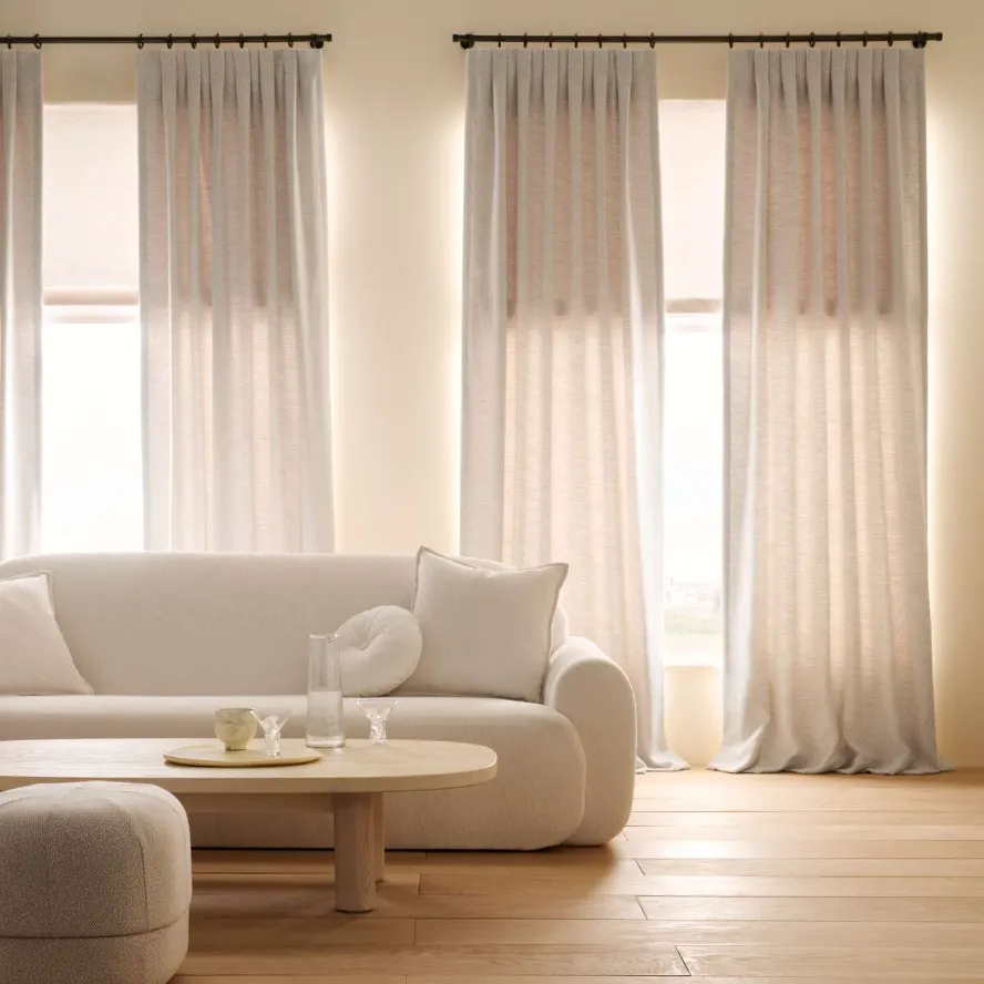 A minimalist living room with a white sofa, round coffee table, and ottoman. Light beige curtains cover three tall windows. Natural light fills the room, highlighting the wooden floor.
