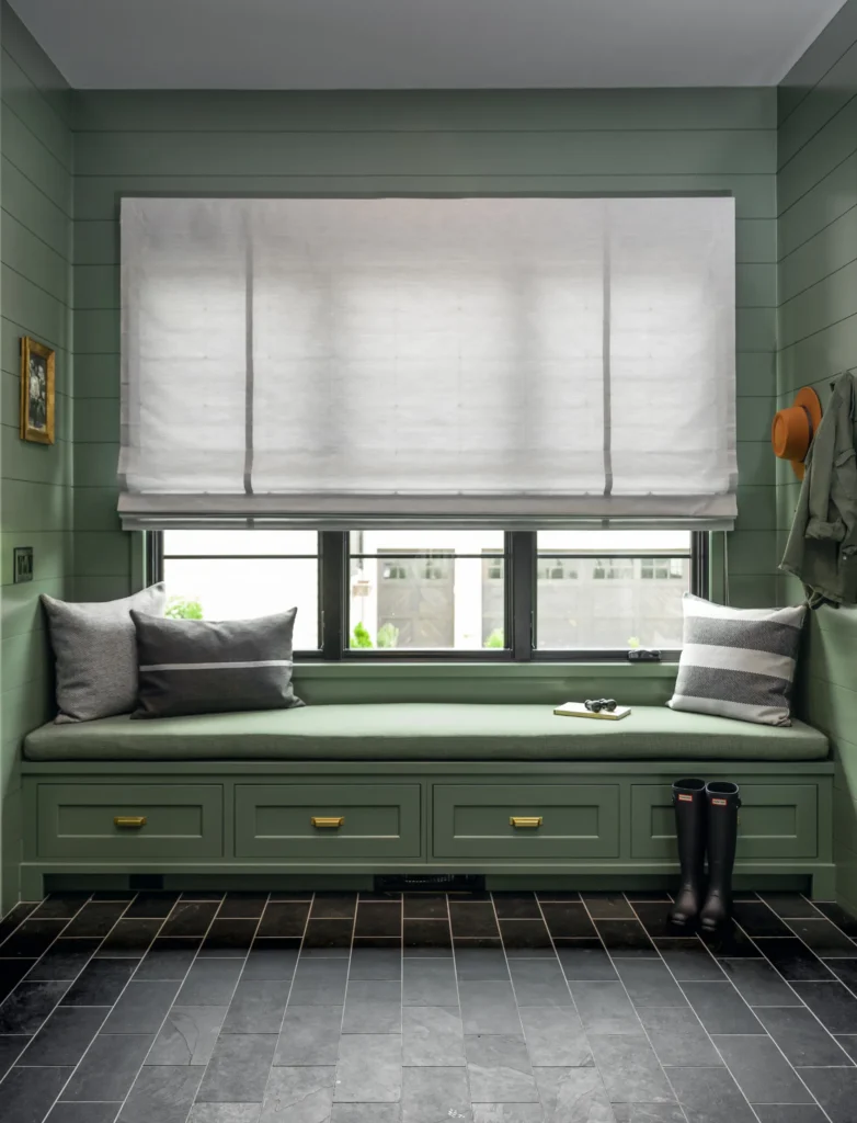 Cozy window nook with a green cushioned bench, pillows, and storage drawers. Large window with a white Roman shade. Dark floor tiles, hat, coat, and boots by the side.