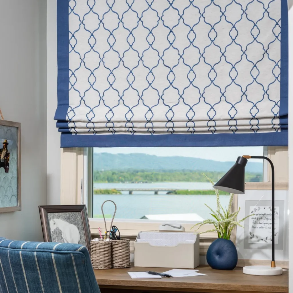A cozy desk setup by a window with a blue and white patterned Roman shade, blue table lamp, plant, and decorative items. The window overlooks a serene lake and hills.