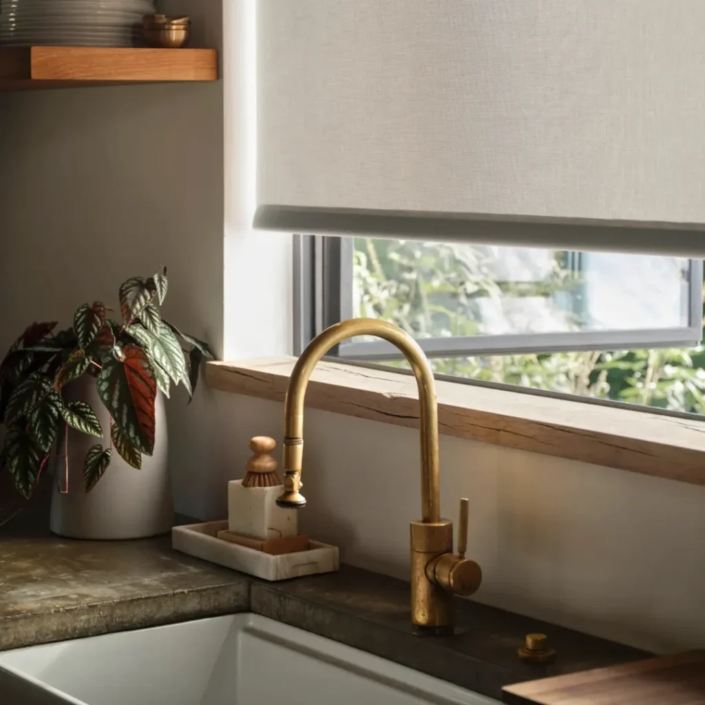 A kitchen sink with a brass faucet, a potted plant, soap, and a window above it with a rolled-up shade.