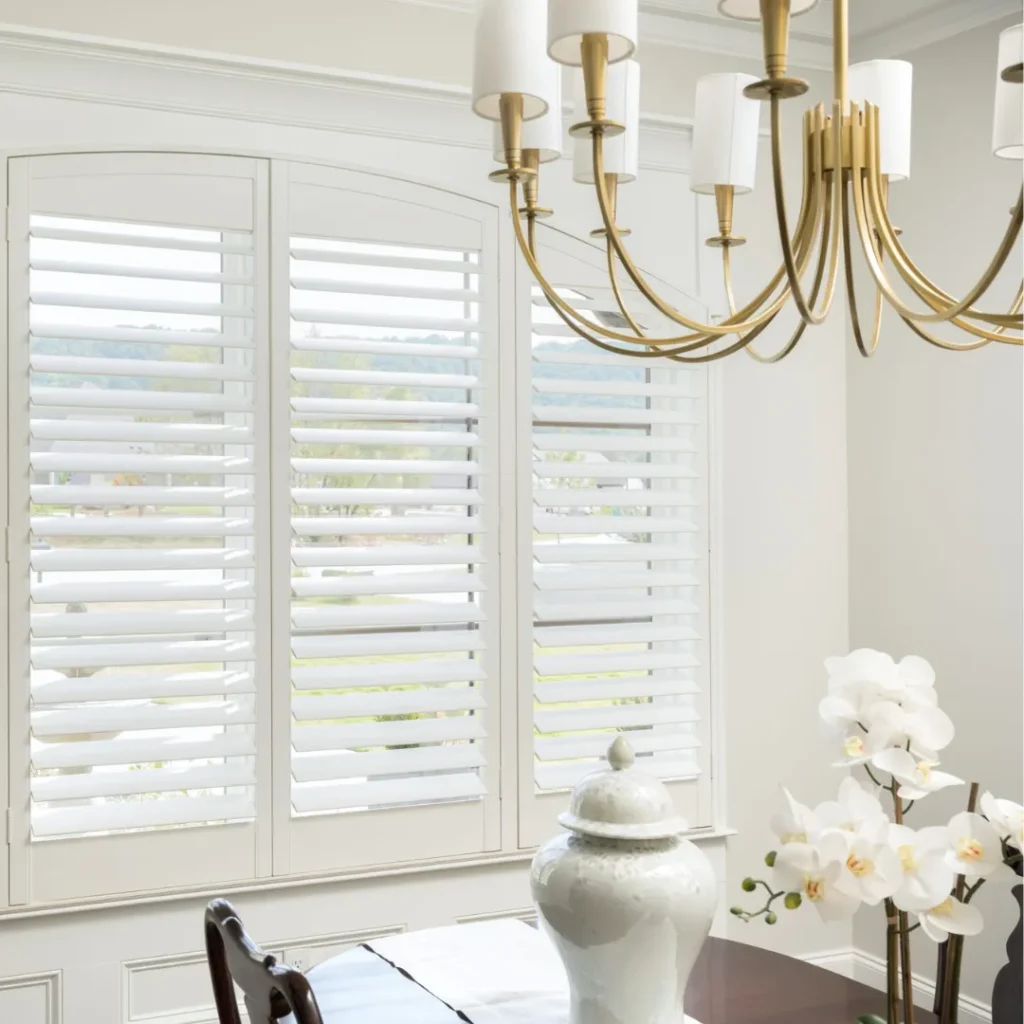 A dining room with white plantation shutters, a brass chandelier, a glossy table, a white jar, and white orchids.