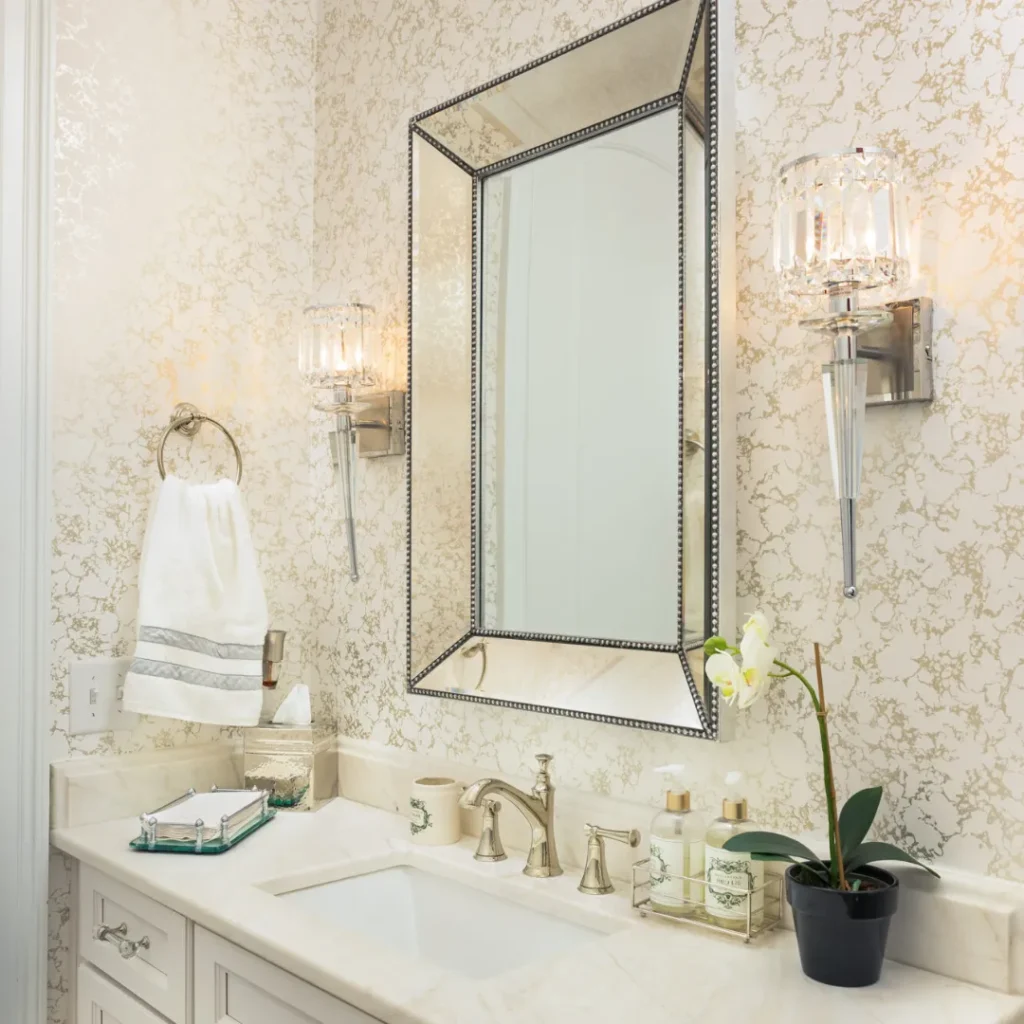 Elegant bathroom with a marble countertop, ornate mirror, and wall-mounted lights. A towel hangs on a ring. A small orchid and toiletries are placed on the sink. Textured wallpaper adorns the walls.