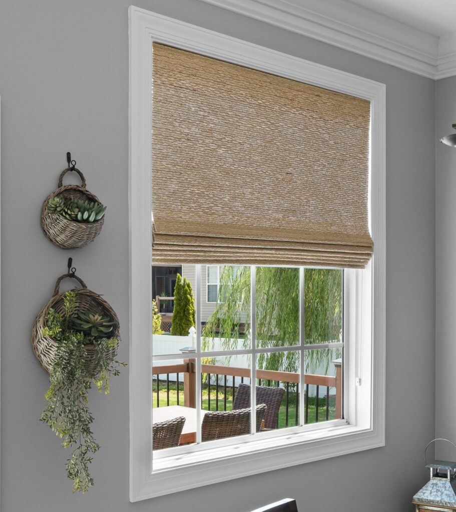 A dining room with a window covered by a woven shade, green plants on the wall, black metal shelves with decor, and a chandelier. A deck and greenery are visible outside.