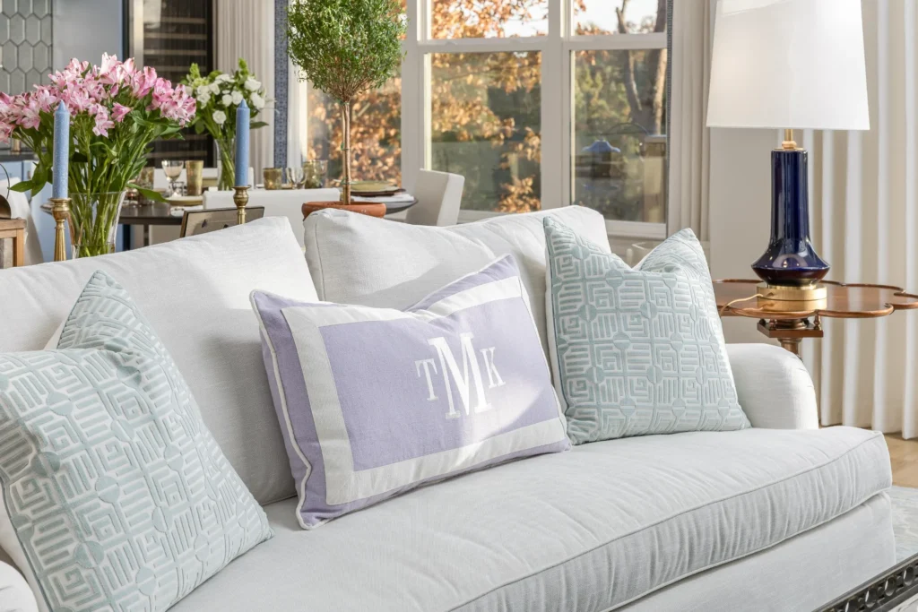 A white sofa with three decorative cushions, one monogrammed, is in a bright living room. A lamp and flowers on side tables are visible, with a view of the outside through large windows.
