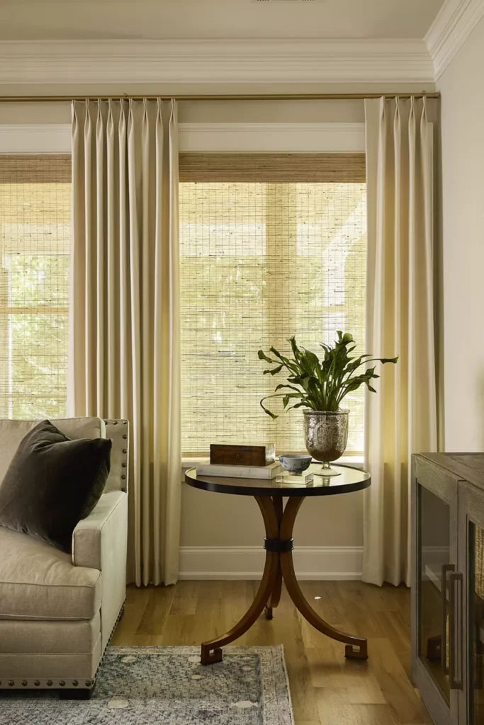 Living room with a beige sofa, round wooden table with a plant, wicker blinds, and long curtains framing the window. A carpet covers part of the wooden floor.