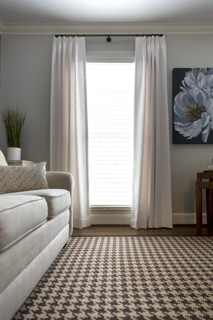 Living room with a beige sofa, white curtains, a black and white patterned rug, a side table, and a large floral painting on the wall.