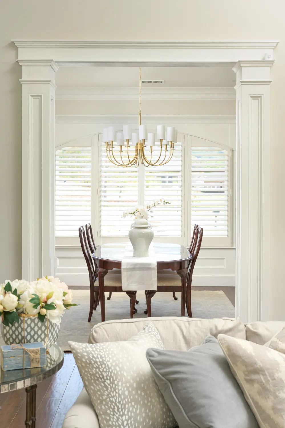 Elegant dining room with a wooden table, chairs, a central white vase, and a gold chandelier. Large windows with shutters are in the background. Soft beige couch in the foreground.
