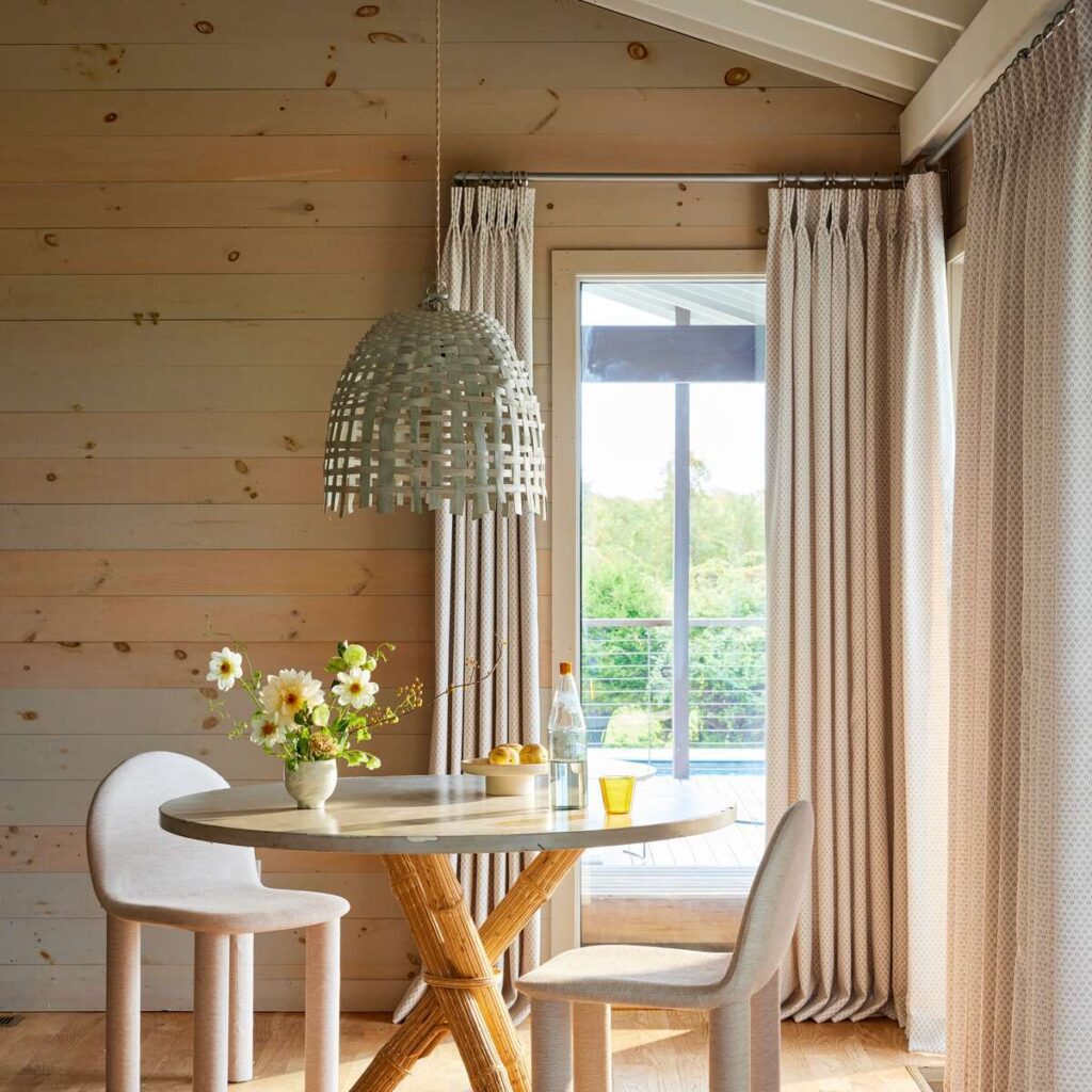 A small dining area with a round table, two chairs, and a woven pendant light. Flowers, fruit, and a glass are on the table. Light wooden walls and curtains frame a window with a view.