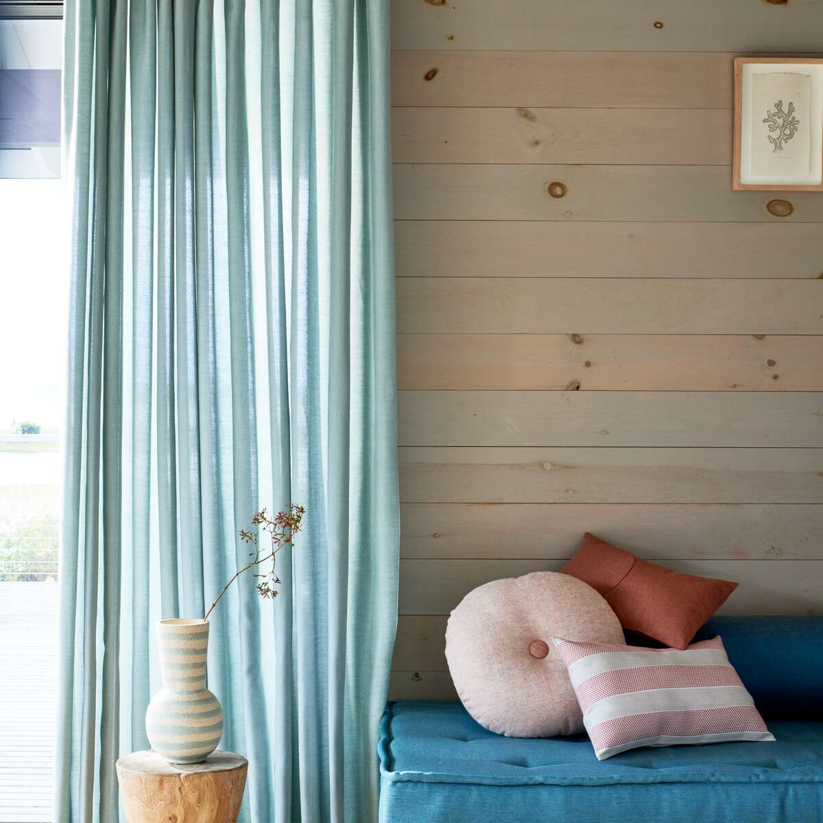 A cozy corner with a cushioned bench and a small vase on a wooden stool beside blue curtains. The wall is wooden with a small framed picture.