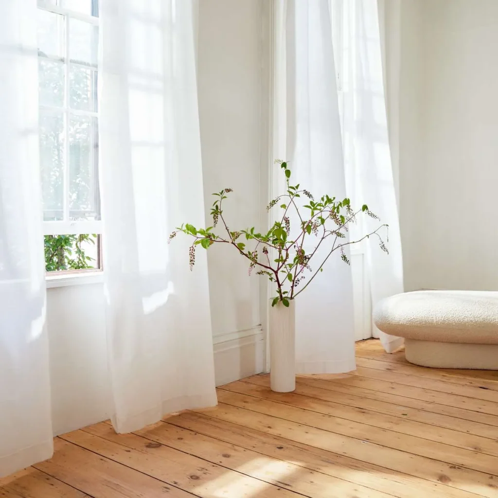 A minimal room with wooden floors, white sheer curtains, a potted plant, and a round, plush cream ottoman. Natural light streams in through the windows.