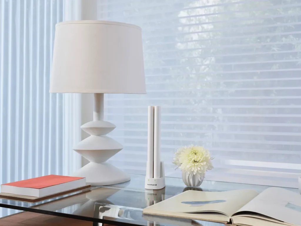 A white modern lamp, a cylindrical device, and a vase with white flowers sit on a glass table next to an open book and stack of closed books. Background shows sheer blinds on a window.