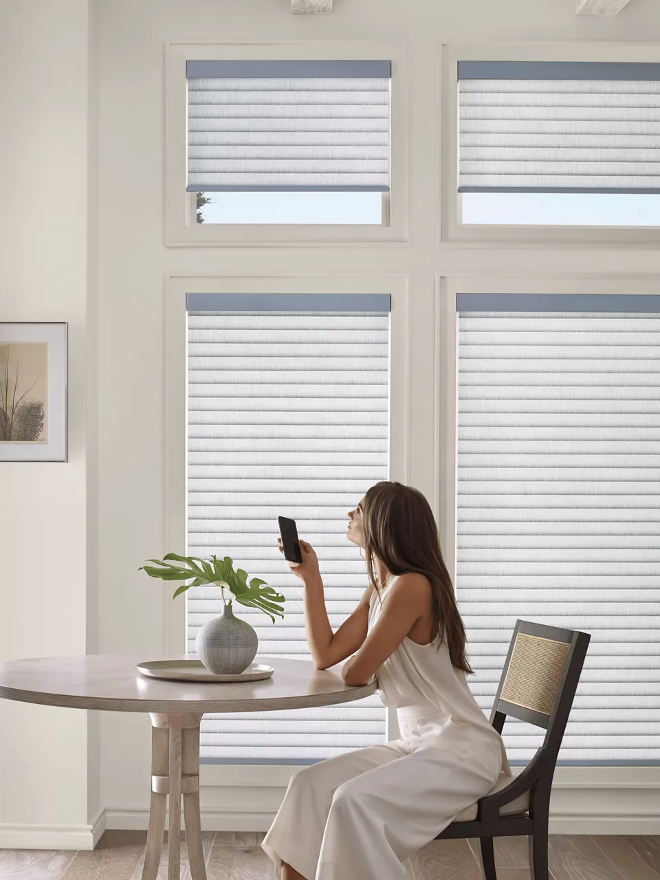 A woman in a white dress sits at a round table, holding a smartphone near her face. There is a potted plant on the table, and large windows with blinds in the background.