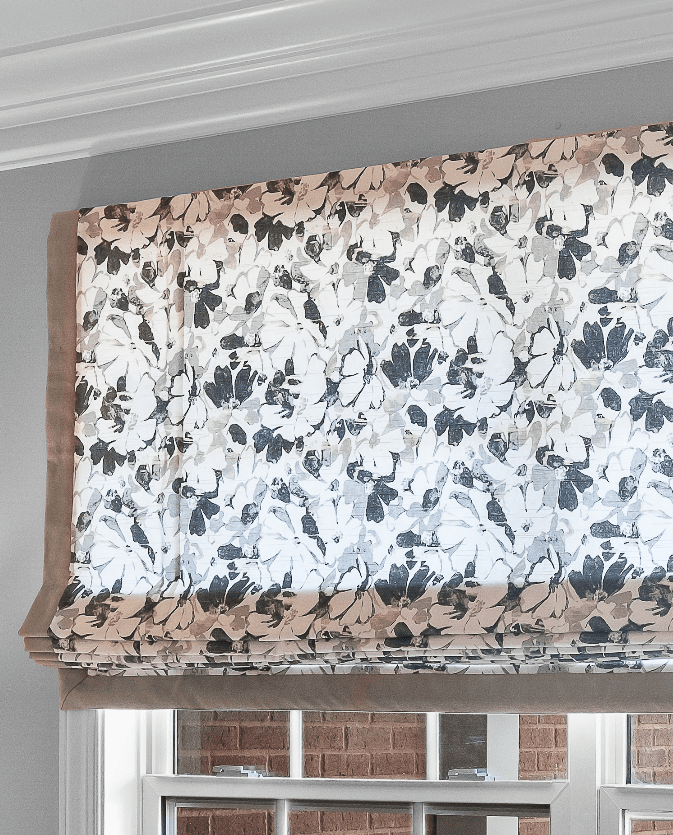 Floral-patterned Roman shade partially open on a window with a white frame, revealing a brick wall outside.