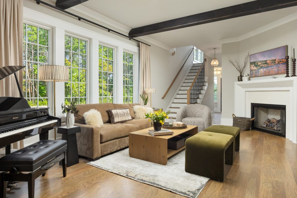 Modern living room with large windows, a brown sofa, a piano, a coffee table, two ottomans, a fireplace, and a staircase in the background.