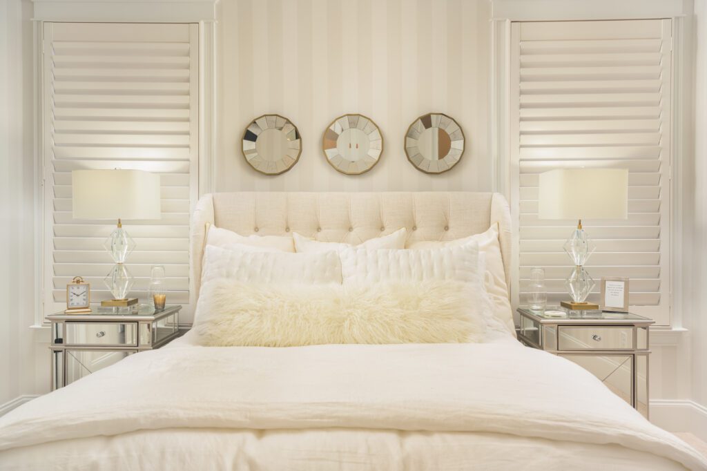 A neatly made bed with white bedding is centered between two mirrored nightstands, each with a lamp. Three round mirrors hang on the striped wall above the bed.