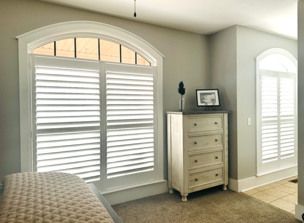 A bright bedroom with large arched windows featuring white shutters, a cream dresser with a framed photo and small decor, and part of a bed with a quilted cover.