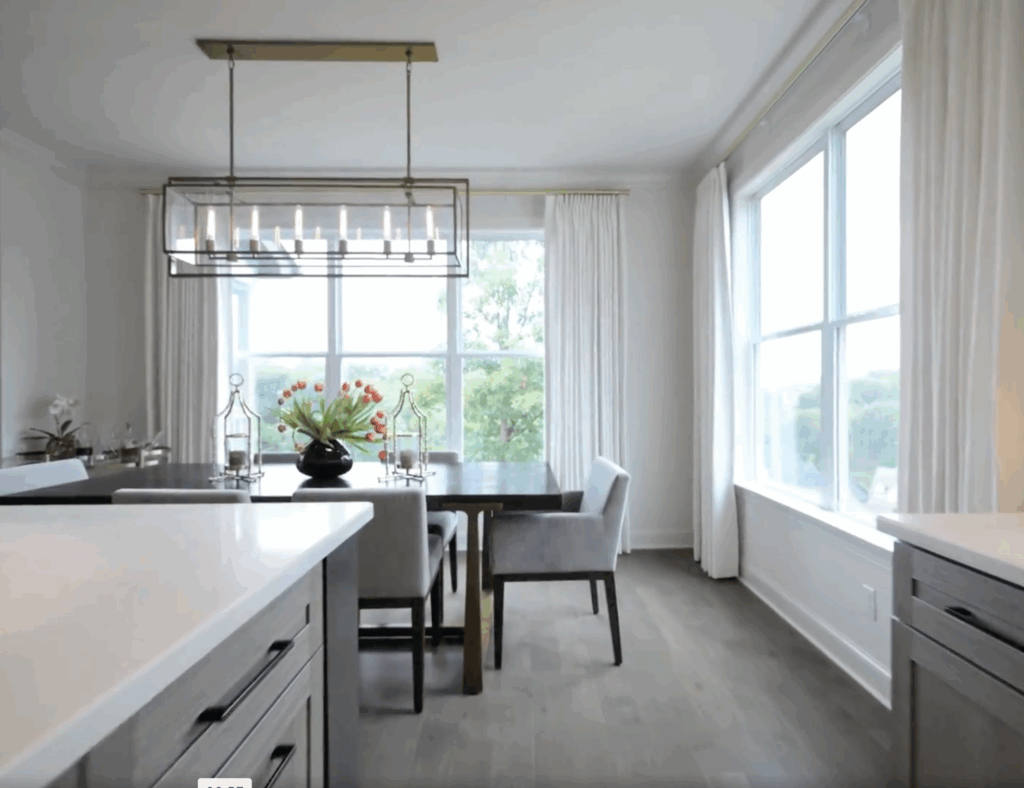 A modern dining room with a dark wooden table, gray upholstered chairs, a large window with white curtains, and a rectangular chandelier overhead.