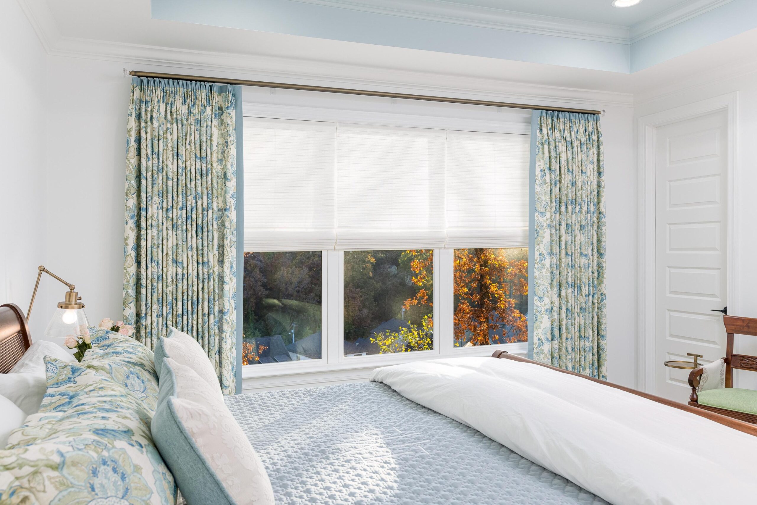 Bright bedroom with a neatly made bed, floral curtains, white blinds, and sunlight streaming through large windows with trees visible outside.