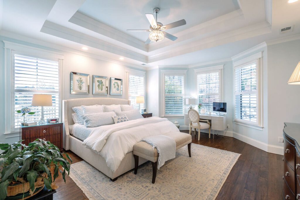 Bright bedroom with a large bed, bench, two bedside tables, a desk with a computer by the window, hardwood floors, and several plants.