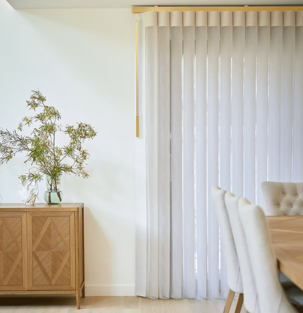 A dining room with a wooden table, upholstered chairs, a sideboard with a vase of greenery, and vertical white blinds covering a large window.