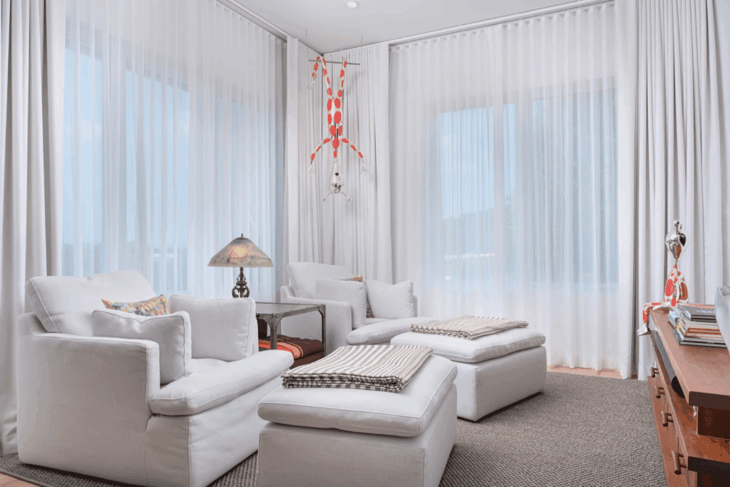 Bright living room with white armchairs, ottomans, a side table with a lamp, sheer curtains, and a red and white hanging mobile.
