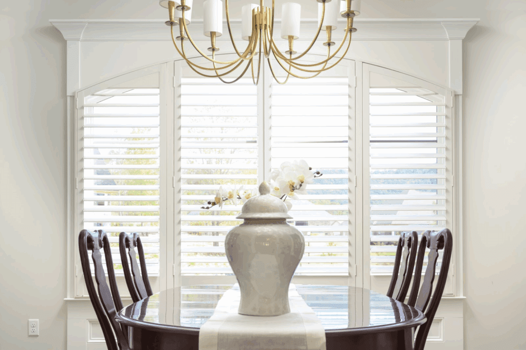 A dining room with a dark wood table, six chairs, a white vase with flowers, a chandelier above, and white shuttered windows in the background.