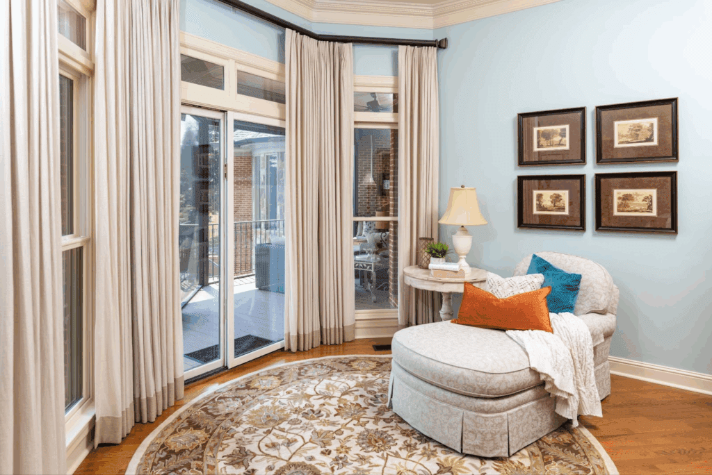 A cozy sitting area with a patterned rug, upholstered chaise lounge, side table with a lamp, and four framed pictures on a light blue wall beside large windows and glass doors.