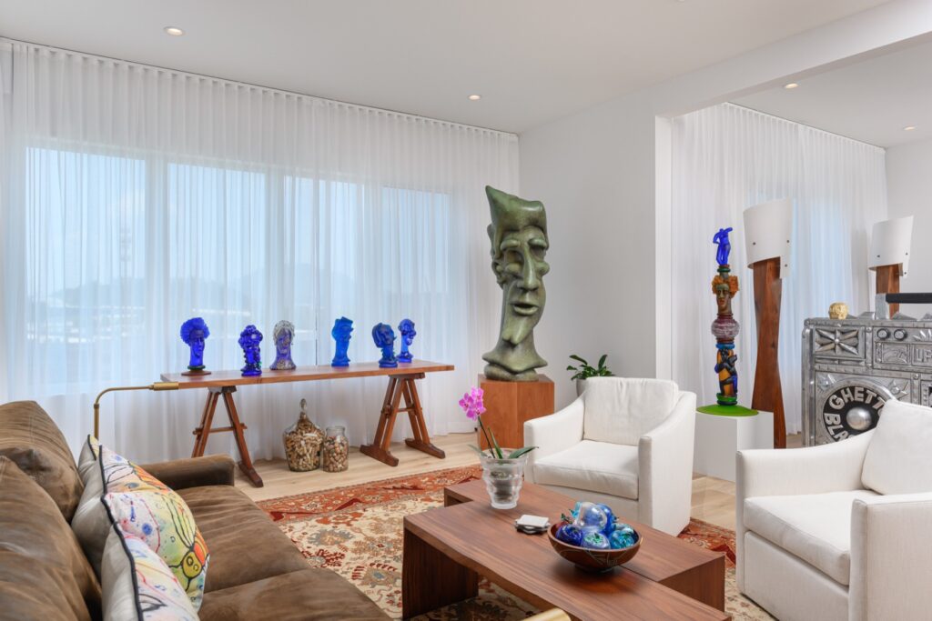 Modern living room with eclectic sculptures, including blue and green abstract heads, a brown leather sofa, two white chairs, and a wooden coffee table on a patterned rug.