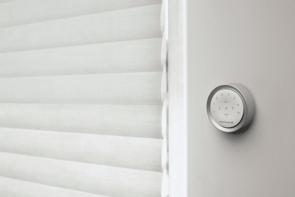 A modern smart wall-mounted control panel is positioned next to a set of closed, white, horizontal window blinds.