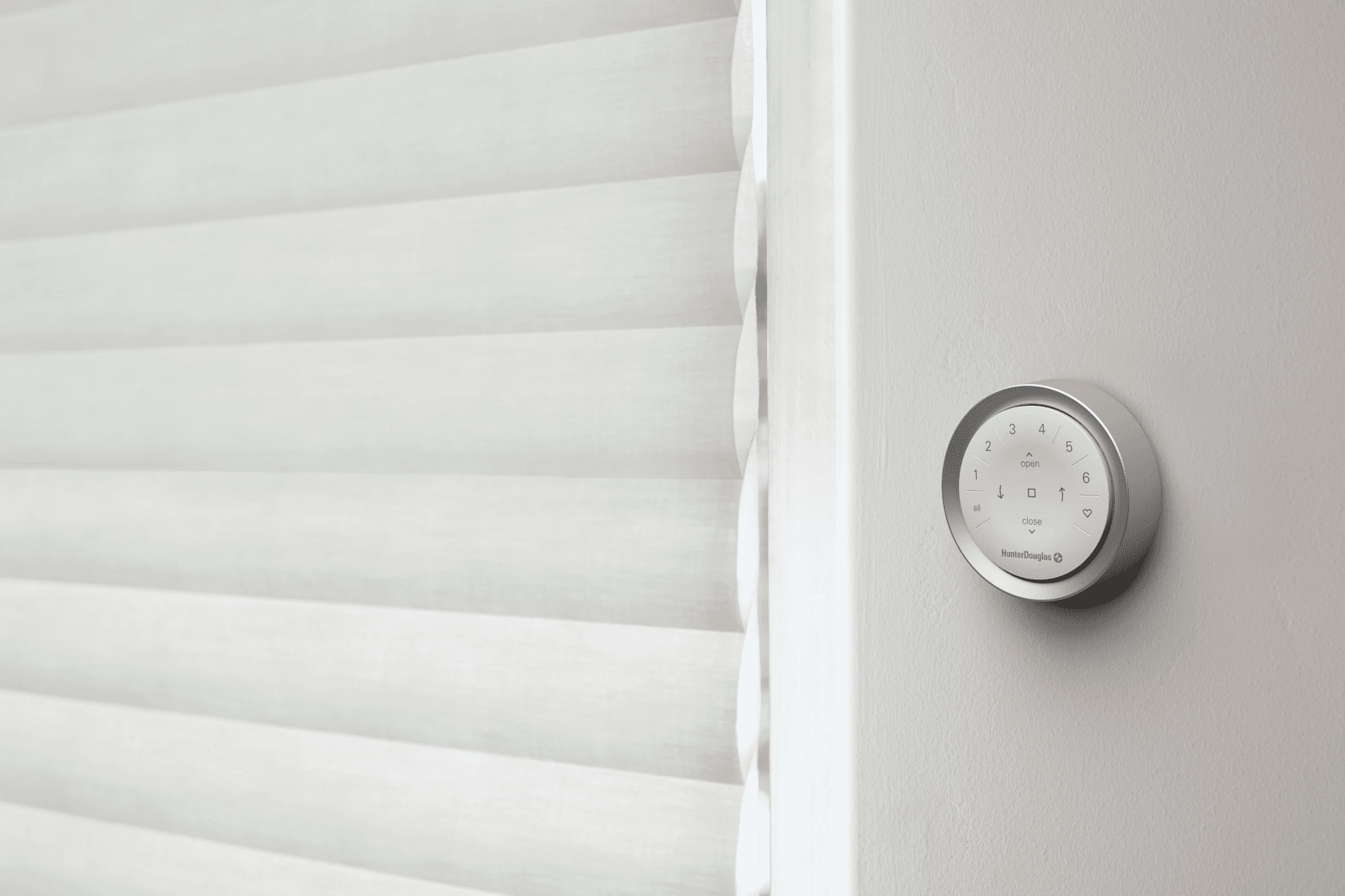 A modern smart wall-mounted control panel is positioned next to a set of closed, white, horizontal window blinds.