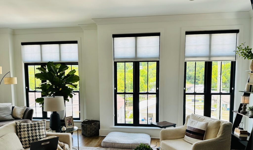 A bright living room with three tall windows, partially covered by white blinds, contains a sofa, armchair, potted plant, and various decor items. Trees and a road are visible outside.