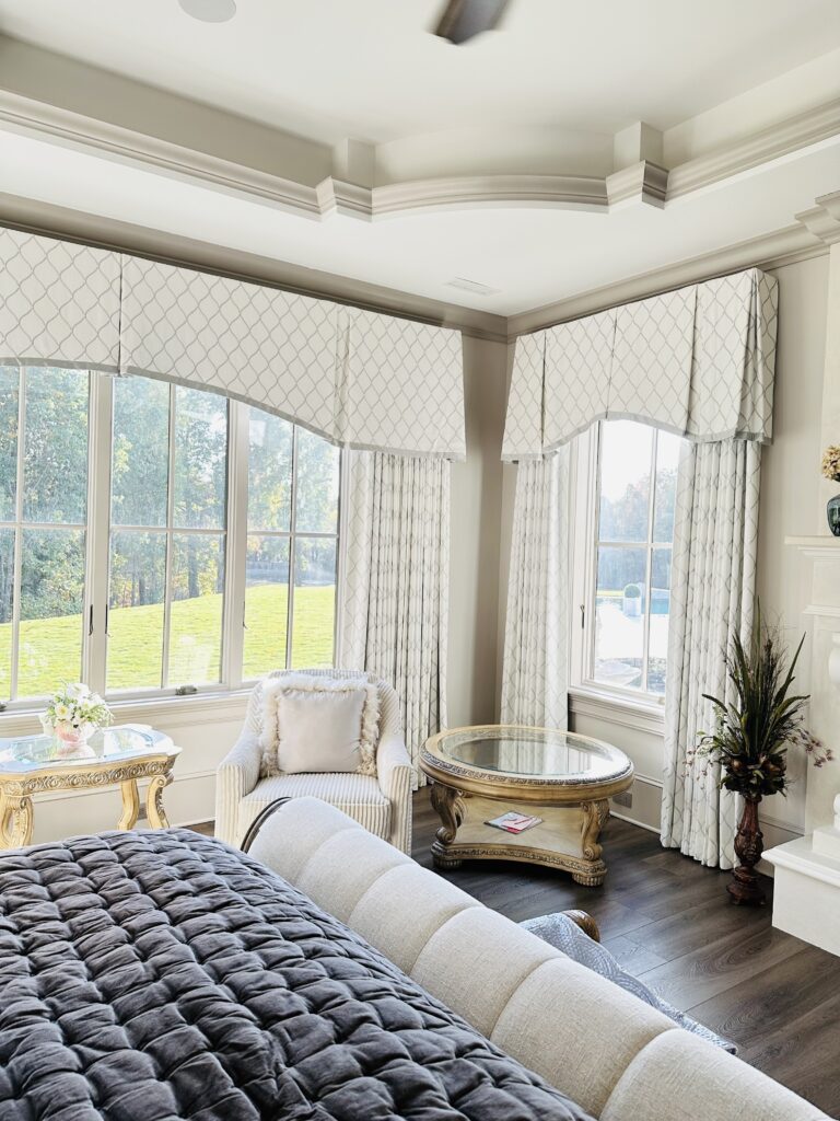 Bright bedroom with large windows, patterned valances and curtains, an armchair, two ornate tables, and a cushioned bed. Sunlight streams in, highlighting neutral decor.
