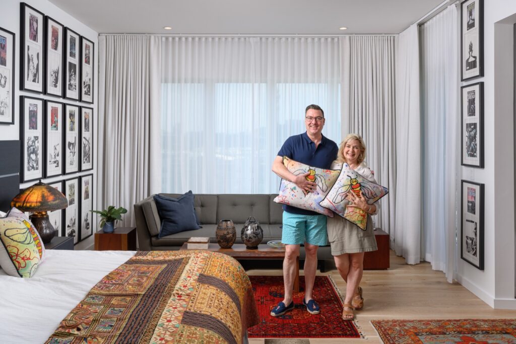 A man and woman stand in a well-decorated bedroom holding colorful pillows, with framed art on the walls and patterned textiles throughout the room.