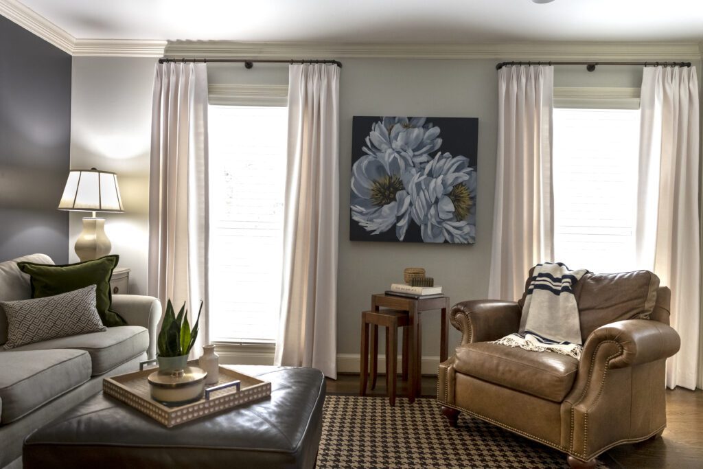 A living room with a beige sofa, brown leather armchair, dark ottoman, and side tables, decorated with a lamp, plants, and a floral painting on the wall between two windows with white curtains.