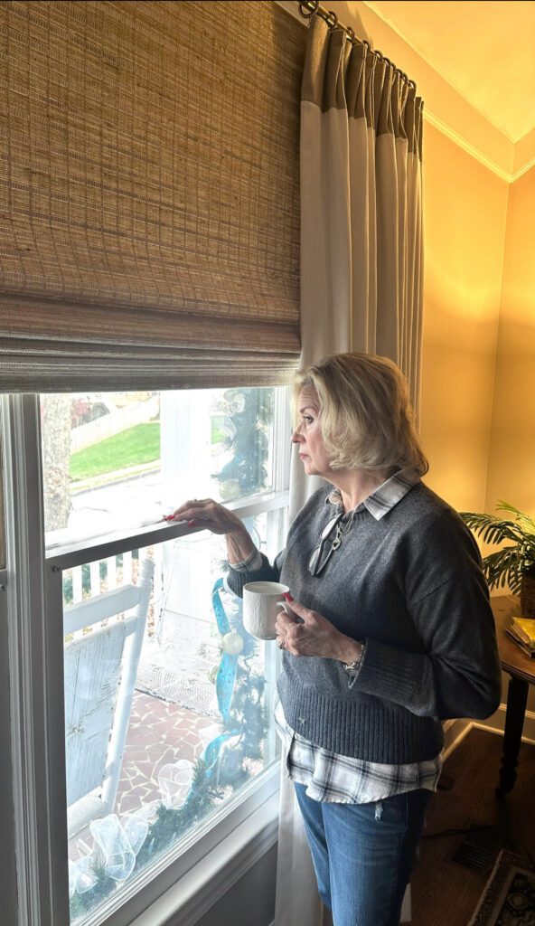 An older woman stands indoors by a window, holding a mug and looking outside with a neutral expression. Daylight illuminates the room.