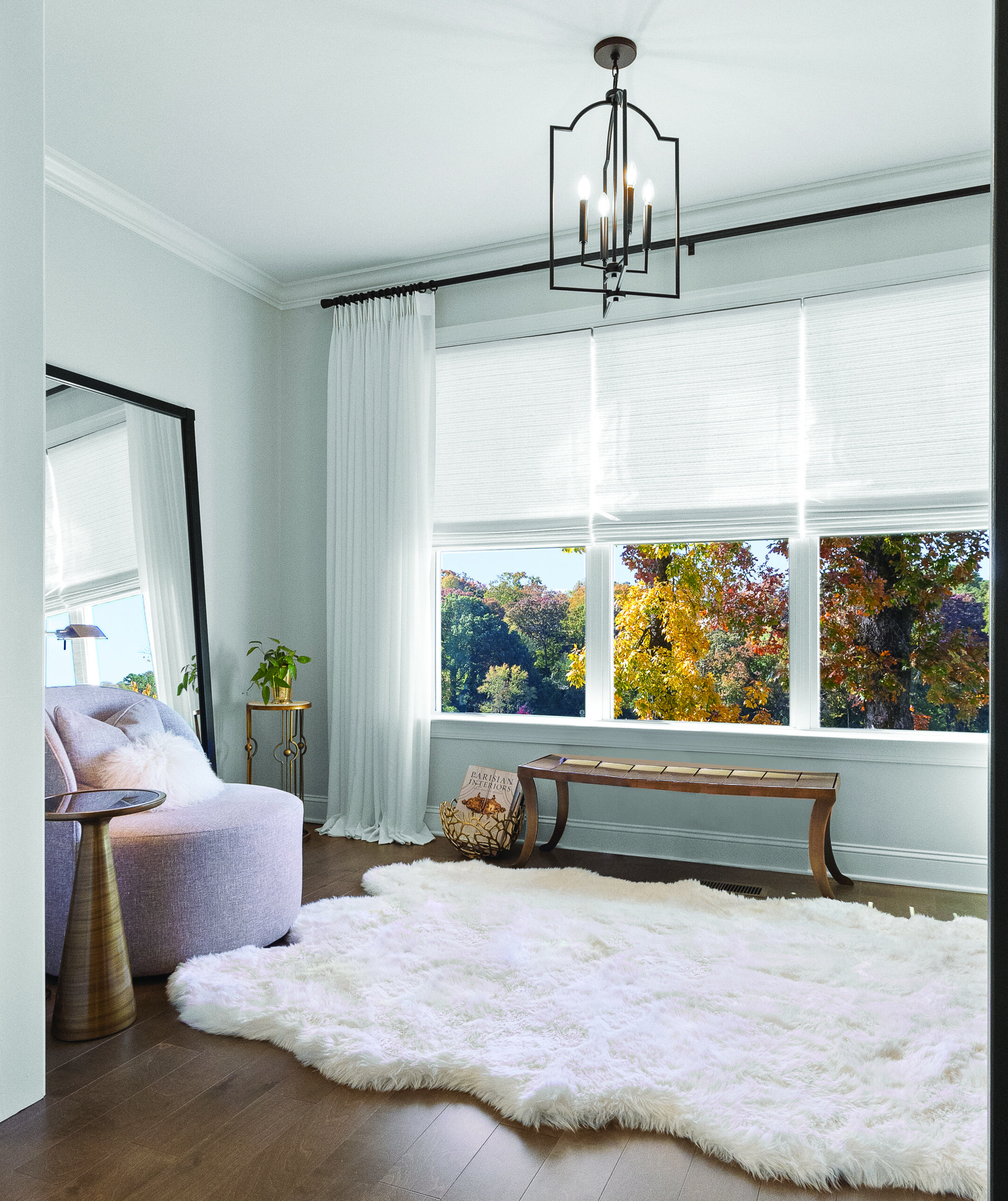 A bright room with large windows, white curtains, a fluffy white rug, a wooden bench, an armchair, a potted plant, and a floor mirror. Autumn trees are visible outside.