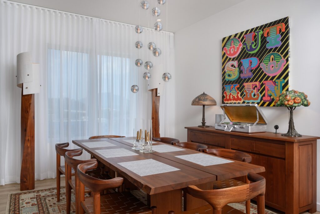 A modern dining room with a wooden table and chairs, pendant lights, a sideboard, decorative objects, and colorful artwork reading "OUTSPOKEN" on the wall.
