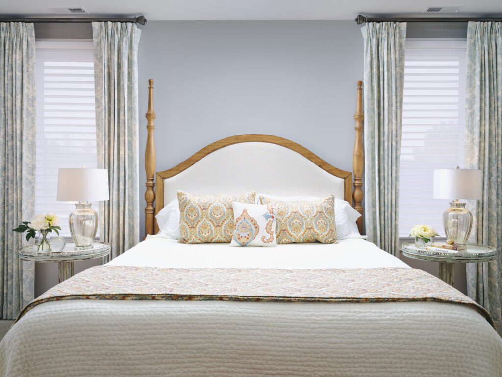 A neatly made bed with a tall upholstered headboard, two patterned pillows, and matching lamps on round bedside tables in a bright bedroom with light-colored curtains.